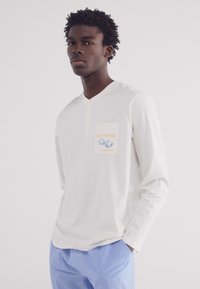 Camicia bianca a maniche lunghe con scollatura a V e dettagli con bottoni, caratterizzata da una tasca anteriore con testo blu e giallo. Abbinata a pantaloni a righe bleu chiaro.