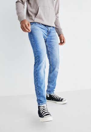 Personne portant un jean slim bleu clair et des baskets montantes noires avec des lacets blancs, debout devant un fond blanc uni.