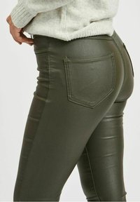 Tummanvihreät keinonahkaleggingsit, joissa on tyylikäs muotoilu, takakappaleeseen sijoitettu tasku ja hienot ompelusaumat, jotka näkyvät lantion alueella.