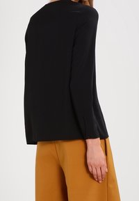 Blusa nera a maniche lunghe con scollatura rotonda, texture liscia, dettagli con bottoni sul retro e vestibilità rilassata, abbinata a pantaloni color senape.
