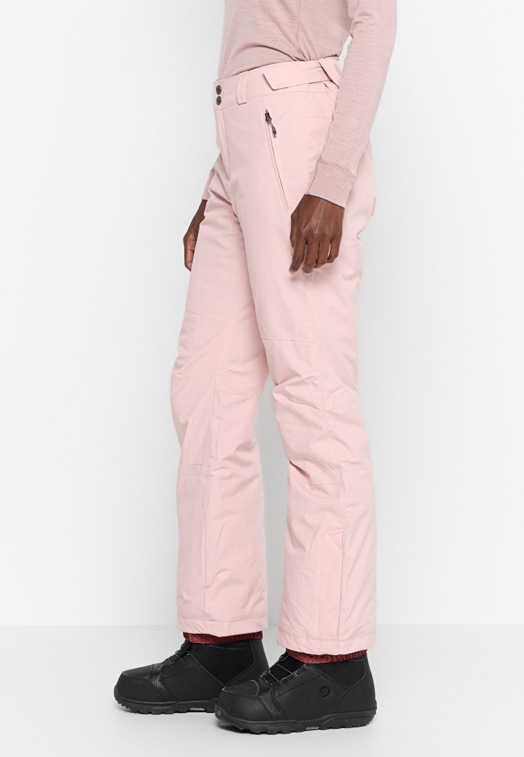 Jasnoróżowe spodnie narciarskie wykonane z wodoodpornego materiału, o prostym kroju, z zamkiem błyskawicznym w kieszeni i wzmocnionymi mankietami, zestawione z czarnymi butami.
