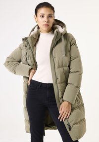 Abrigo tipo puff en verde oliva con capucha, cierre frontal de cremallera, bolsillos laterales y un forro suave. Llevado sobre un suéter de canalé de color claro y jeans oscuros.