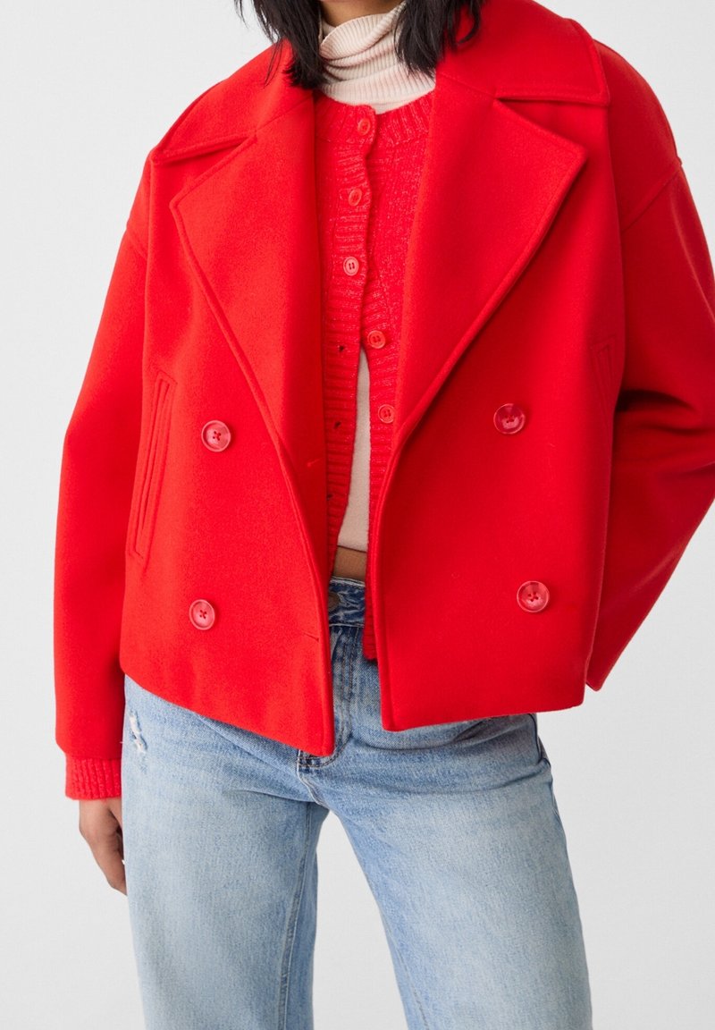 Personne portant un manteau rouge vif croisé par-dessus un cardigan rouge et un col roulé clair, associé à un jean bleu clair.