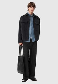 Zwarte corduroy jas met twee borstzakken, gelaagd over een licht denim shirt, gecombineerd met zwarte wijde jeans en een zwarte draagtas.