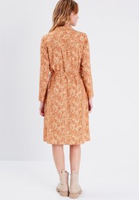 Langärmliges Kleid in Orange mit floralem Muster, ausgestattet mit einem taillierten Bund und knielangem Saum. Kombiniert mit beigen Ankle Boots.