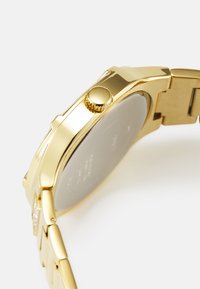 Ceas de aur cu un carcasă metalică netedă, coroana texturată și o brățară cu verigi. Prezintă un design minimalist și un finisaj lustruit.