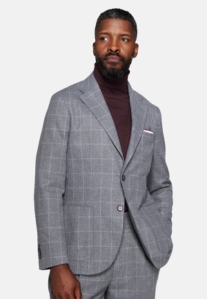 Man with a beard wearing a gray checkered suit and dark turtleneck, standing with one hand in his pocket, looking slightly to the side.