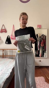 Šedé široké tepláky s elastickým pasem, modelka drží transparentní balení označené "WEEKDAY," které předvádí měkkou strukturu látky.