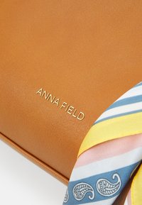 Hellbraune Handtasche aus Kunstleder mit einem goldgeprägten Logo, verziert mit einem bunten, gemusterten Seidenschal in Blau-, Gelb- und Rosatönen.