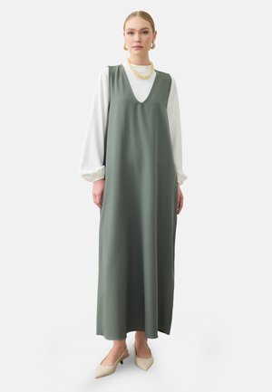 Donna che indossa un lungo abito verde salvia senza maniche sopra un top bianco con maniche a sbuffo, scarpe beige con punta, orecchini a cerchio dorati e una collana massiccia, in piedi dritta.