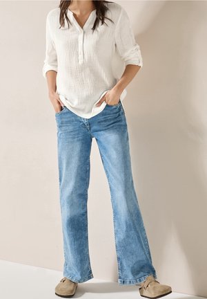 Kvinde iført hvid tekstureret langærmet skjorte med op-rullede ærmekanter, lyseblå jeans med vide ben og beige slip-on sko, stående med hænderne i lommerne.