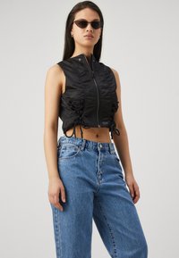 Chaleco negro corto con detalles recogidos y una cremallera central. Llevado sobre unos vaqueros de tiro alto en azul claro. Lleva gafas de sol y tiene una textura suave.
