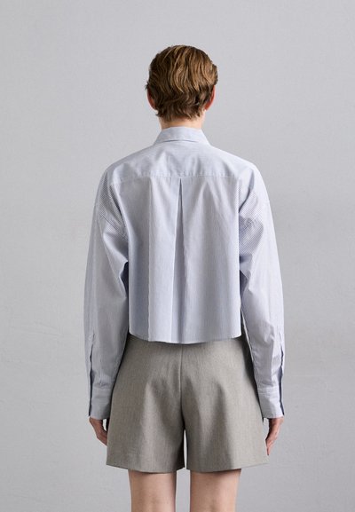 Cropped button-up shirt in light blue and white stripes, with long sleeves and ventilation pleats, paired with gray tailored shorts.