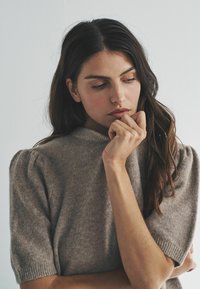 Młoda kobieta z długimi ciemnymi włosami, ubrana w krótkim rękawie szary sweter, patrząca zamyślona w dół, z dłonią blisko twarzy na jednolitym tle.