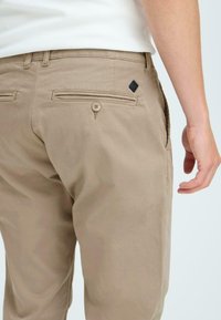Beige cotton trousers with a slim fit, featuring back pockets, button details, and a small black logo patch. Smooth texture.