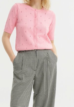Femme portant un pull rose à manches courtes boutonné avec de petits trous et un pantalon à motif pied-de-poule noir et blanc, les mains dans les poches.