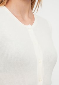 Cardigan alb cu reieuri și decolteu rotund, având patru nasturi albi pe partea frontală. Textura materialului este ușoară și moale.