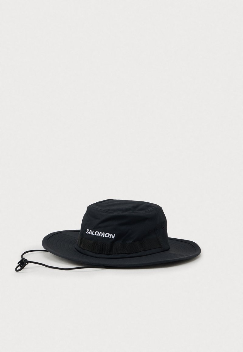 Chapéu Salomon outdoor de aba larga e preta, com fita ajustável no queixo e ilhós de ventilação em tecido, sobre um fundo simples.