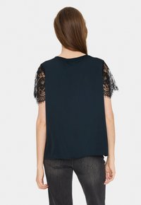 Top blu navy con maniche corte in pizzo. La parte posteriore è liscia con una vestibilità casual, e il collo è rotondo. Indossato con jeans scuri.