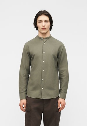 Joven con camiseta verde oliva de cuello mandarín y cierre de botones, y pantalones cargo marrón oscuro, sobre un fondo liso.