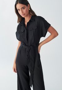Juodas kombinezonas trumpomis rankovėmis, priekyje užsegamas mygtukais, su dviem krūtinės kišenėmis ir surišamu juosmens diržu. Lygios tekstūros audinys, laisvas kirpimas.