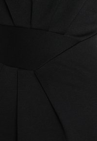 Černé šaty s texturovanou látkou, šikmými detaily a širokým hladkým pasem, které zdůrazňují strukturovaný design.