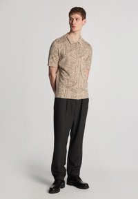 Camicia beige a maniche corte con motivi geometrici in rilievo, abbinata a pantaloni scuri e scarpe nere, in piedi davanti a uno sfondo neutro.