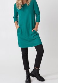 Teal tuniek jurk met driekwart mouwen, twee zijzakken, gedragen met zwarte slim-fit broek en zwarte veterschoenen. Glad stoffen textuur.