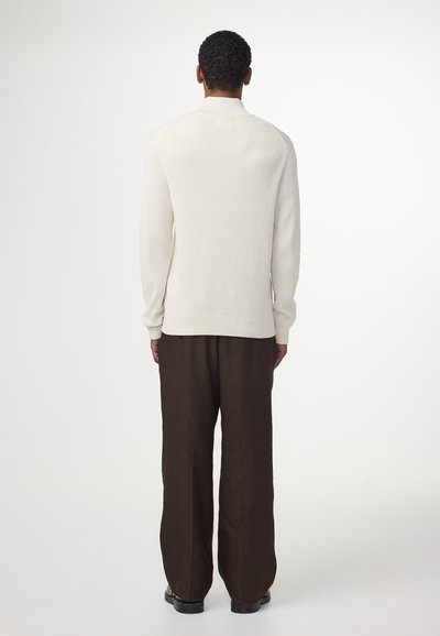 Hellbeige gerippten Pullover mit hohem Kragen, schlanker Passform und langen Ärmeln, kombiniert mit locker sitzenden braunen Hosen und schwarzen Schuhen.