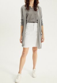 Kvinde iført grå cardigan og top med blomsterdetaljer, hvid nederdel og hvide sneakers, stående på en lys baggrund.