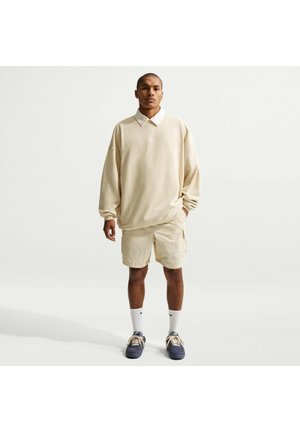 Mand står frontalt iført en beige oversized sweatshirt, beige shorts, hvide sokker og marineblå sneakers mod en ensfarvet baggrund.