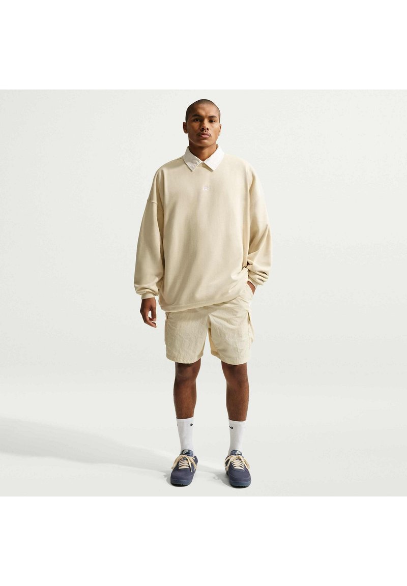 Homme debout face à l'appareil, portant un sweat beige oversize, un short beige, des chaussettes blanches et des baskets bleu marine sur un fond uni.