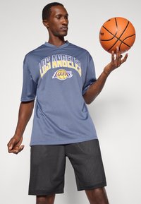 Grå mesh baskettröja med "Los Angeles Lakers" text, tillsammans med svarta shorts med strukturerad mönstring. Spelaren håller en basketboll.