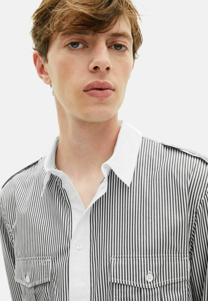 Giovane uomo con capelli castano chiaro che indossa una camicia a maniche lunghe a righe verticali bianco e nere con colletto bianco e taschini sul petto.