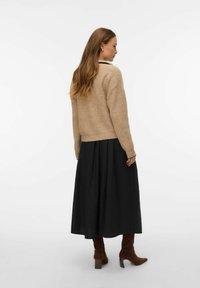 Beżowy sweter z dzianiny z okrągłym dołem, zestawiony z długą czarną spódnicą. Modelka ma na sobie brązowe zamszowe botki, widziana od tyłu.