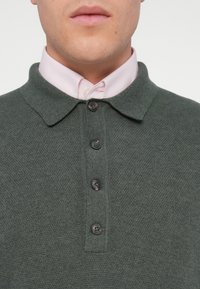 Polo en maille vert foncé avec une finition texturée, doté d'un col, de six boutons noirs et d'une chemise rose clair en dessous.