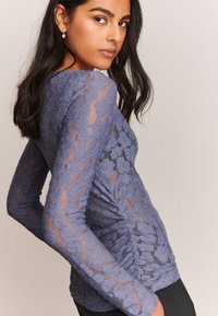 Top in pizzo a maniche lunghe in lavanda tenue, con motivo floreale, dettaglio arricciato ai lati e silhouette aderente. Adatto per essere indossato a strati.