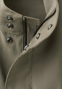 Gros plan d'un tissu vert olive avec une fermeture éclair partiellement ouverte et quatre boutons-pression métalliques sur un vêtement.