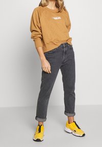 Brun sweatshirt med vit logotyp, grå jeans med uppvikta ärmar och klara gula slipskor med svarta detaljer. Slät tyg, avslappnad passform.