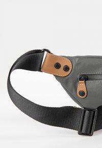 Ce fanny pack en tissu gris est agrémenté de détails en cuir marron, d'une poche zippée, d'une sangle noire et de fermetures à bouton-pression. Fini texturé. Sangle réglable.