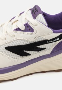 Sneaker mit einem Obermaterial aus Netzstoff in Creme mit lila und schwarzen Akzenten. Dicke Sohle mit strukturierter Oberfläche, mit der Aufschrift "SHADOW" in Weiß.