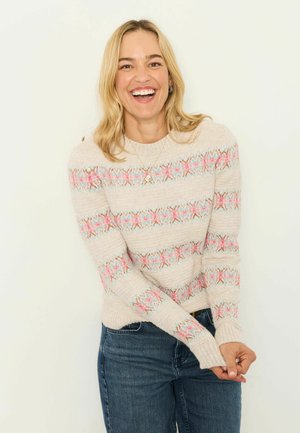 Smiling woman with blonde hair wearing a light beige sweater with pink and blue patterns and blue jeans against a plain white background.
