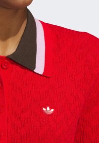 Polo shirt en tricot rouge avec un motif texturé. Présente un col marron avec des rayures roses et blanches et un petit logo Adidas blanc sur la poitrine.