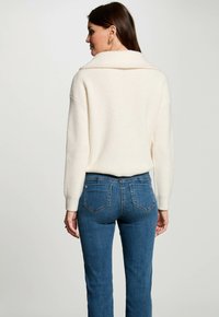 Elfenbeinfarbener, gerippter Pullover mit breitem Kragen, kombiniert mit blauen Jeans im klassischen Schnitt und mit Taschen auf der Rückseite. Gesamt glatte Textur.