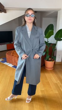 Blazer gris de gran tamaño con solapas anchas, cierre de un solo botón y un bolsillo en el pecho. Lleva puesto sobre una camisa a rayas y combinado con pantalones azul marino y tacones blancos.