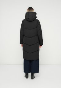 Person står med ryggen til iført en lang sort dunjakke med hætte, brede mørkeblå jeans og sorte ankelstøvler med hæl.