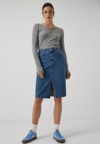 Un pull gris à manches longues avec des boutons associé à une jupe crayon en denim bleu avec une fente à l'avant, porté avec des baskets bleues et des chaussettes blanches.