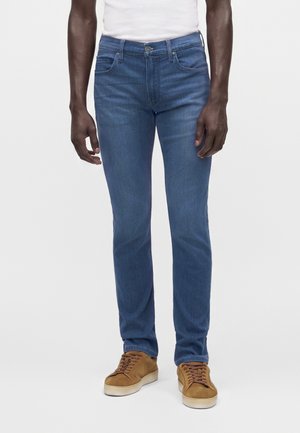 Homme portant un jean slim bleu, une chemise blanche et des baskets marron décontractées, debout devant un fond uni.