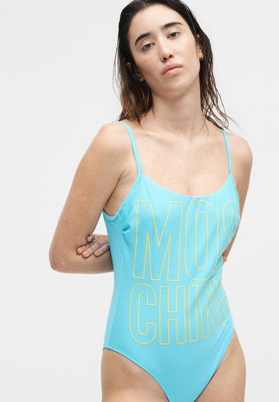 Γυναίκα με βρεγμένα μαλλιά που φοράει ένα ανοιχτό μπλε μαγιό με λεπτές τιράντες και μεγάλο κίτρινο κείμενο "MOSCHINO", στέκεται με τα χέρια πίσω από την πλάτη.