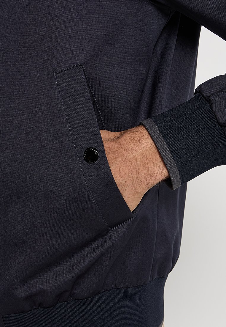 Marineblauer Bomberjacke mit strukturiertem Finish, gerippten Strickbündchen, seitlichem Reißverschlussfach und einem schwarzen Druckknopfakzent.